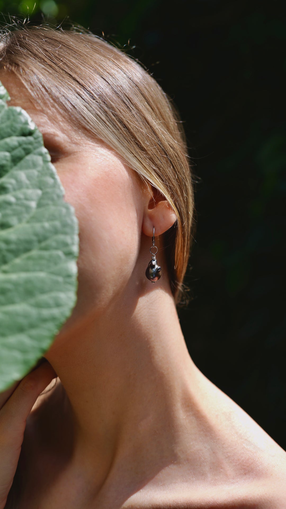 Náušnice z chirurgickej ocele s tmavými sladkovodnými barokovými perlami na okrúhlych mechanických háčikoch. Zobrazené na uchu modelky.