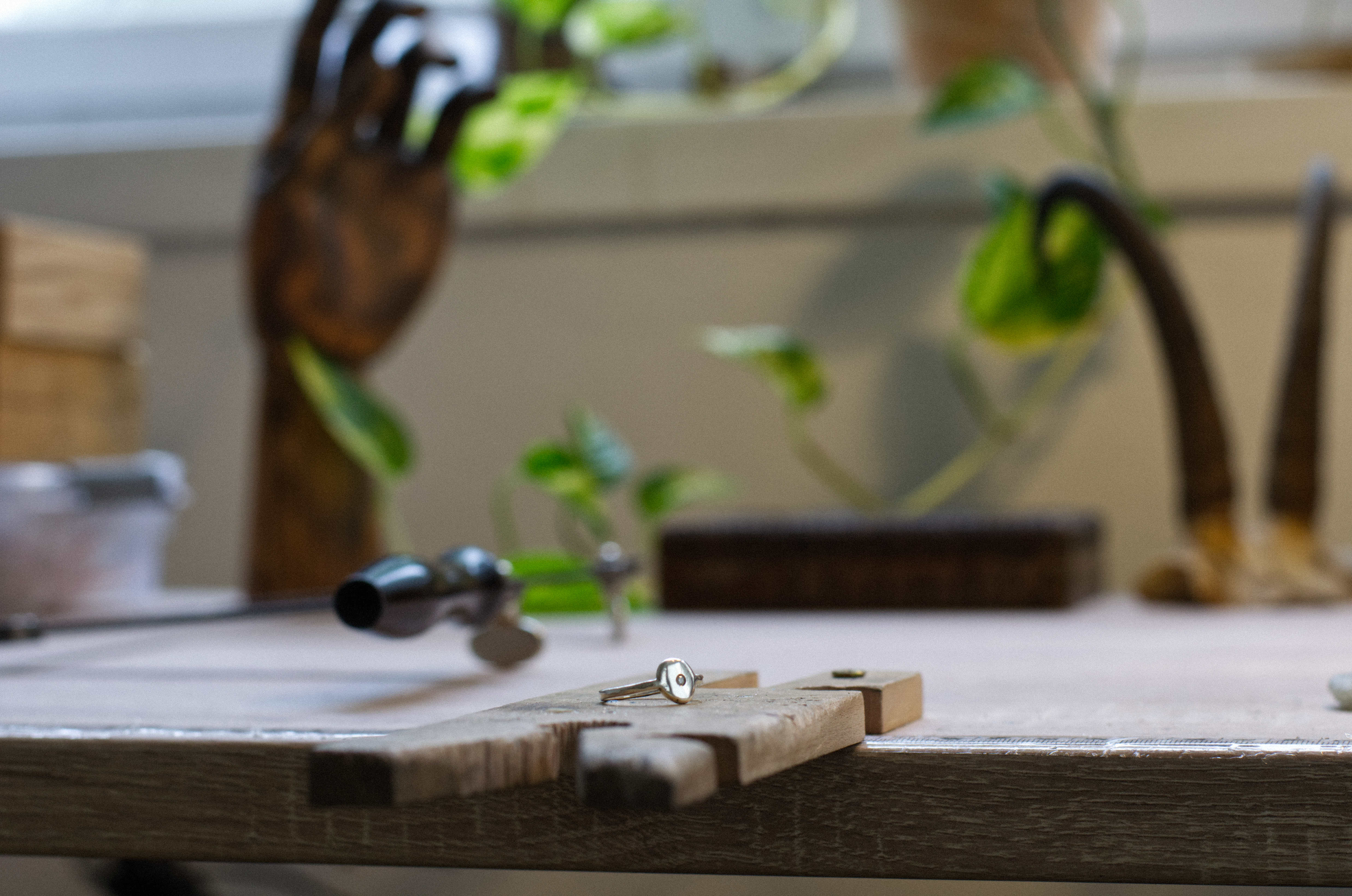 detail on a jewelry making bench pin with a dainty silver ring placed on it. Greenery and vintage decor out of focus in the background